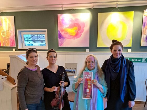 Eine Gruppe von vier Frauen steht in einer Kunstgalerie. Im Hintergrund sind bunte, abstrakte Gemälde zu sehen.