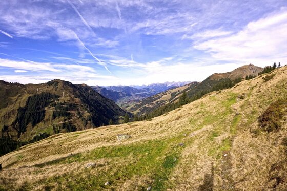 Blick von einem grasbewachsenen Berghang in ein weites Tal unter blauem Himmel mit Kondensstreifen.
