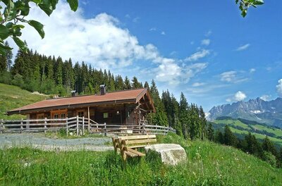 Chalet Wirtsalm Holzhütte mit Zaun auf einer grünen Wiese vor Wald und Gebirgskette unter blauem Himmel.