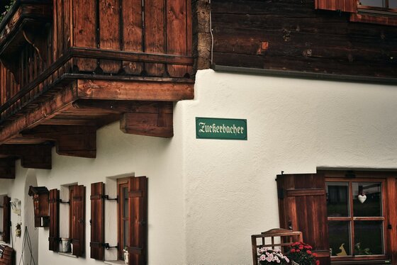 Fassade des Astlingerhofs Nahaufnahme einer Hauswand mit Holzfensterläden, Balkon und einem grünen Schild mit der Aufschrift Zuckerbacher.