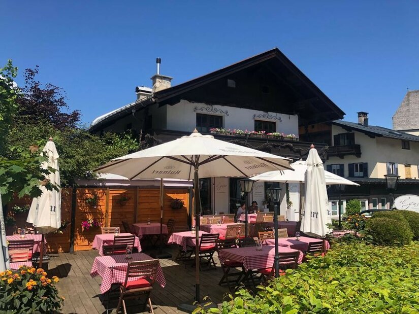 Il Gusto Sommer Außenterrasse eines Restaurants mit rot-weiß karierten Tischdecken und weißen Sonnenschirmen vor einem Alpenhaus.