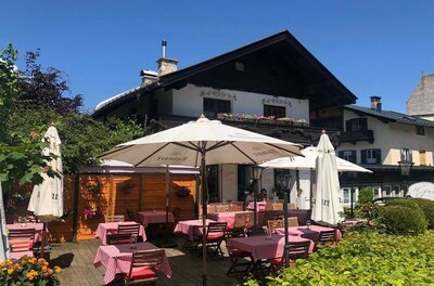 Außenterrasse eines Restaurants mit rot-weiß karierten Tischdecken und weißen Sonnenschirmen vor einem Alpenhaus.