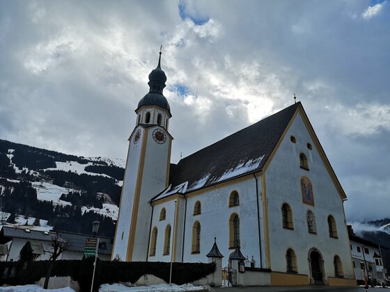 Weiße Kirche mit Zwiebelturm vor bewaldeten, schneebedeckten Bergen unter bewölktem Himmel.