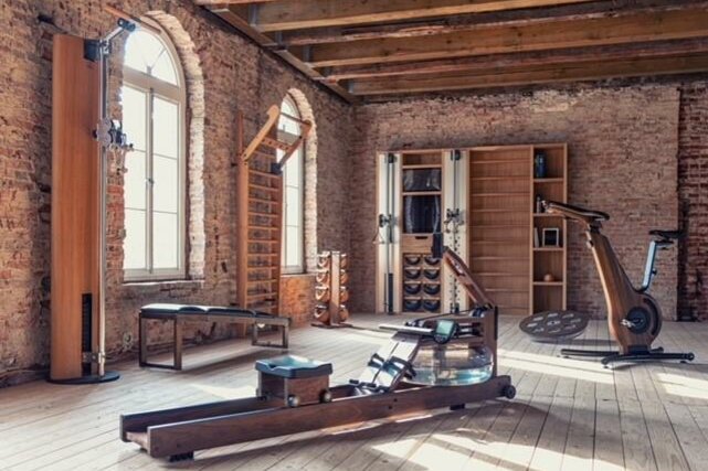 Fitnessraum mit Holzgeräten, Rudermaschine und Ergometer vor einer Ziegelwand.