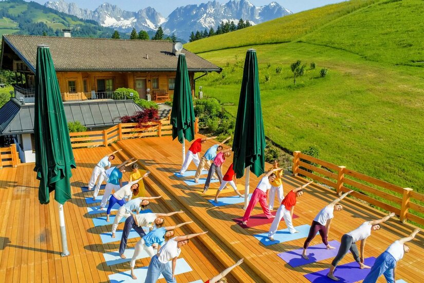 Eine Yoga-Gruppe praktiziert im Freien auf einer Holzterrasse mit Blick auf die Berge. Der Himmel ist klar und es ist sonnig.