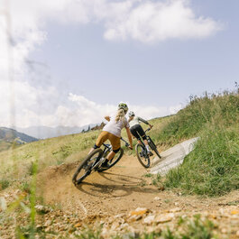 Sonnenrast Trail in Kitzbühel: two mountain bikers ride through curves on a green trail under a blue sky.