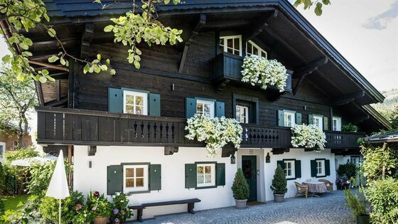 Das historische Weberhaus Zweistöckiges Alpenhaus mit dunkler Holzfassade, weißen Wänden im Erdgeschoss und blumengeschmückten Balkonen.