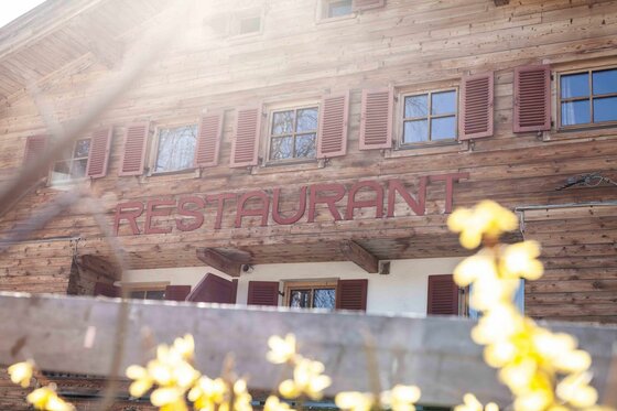 Restaurantfassade des Bruggerhofs Holzfassade eines alpinen Gebäudes mit dem Schriftzug Restaurant und roten Fensterläden.