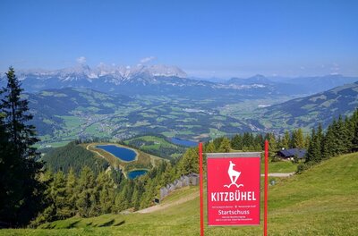 Panoramablick am Hahnenkamm Blick vom Hahnenkamm auf Kitzbühel mit rotem Startschuss-Schild, Speicherseen und Gebirgskette im Hintergrund.
