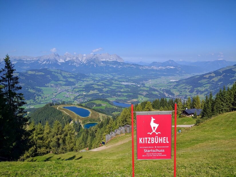 Blick vom Hahnenkamm auf Kitzbühel mit rotem Startschuss-Schild, Speicherseen und Gebirgskette im Hintergrund.
