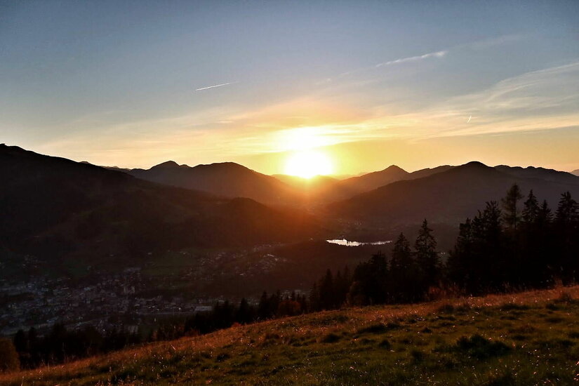 Blick von der Mittelstation Sonnenuntergang über einer bergigen Landschaft mit einem Tal und Nadelbäumen im Vordergrund.