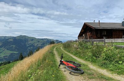Mountainbike am Panoramaweg Ein rotes Mountainbike liegt auf einem Schotterweg neben einer Holzhütte vor Bergpanorama.