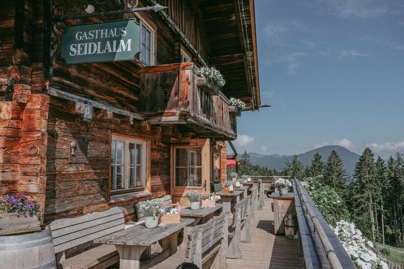 Holzfassade des Gasthauses Seidlalm mit Außenterrasse, Holztischen und Blick auf bewaldete Berge.