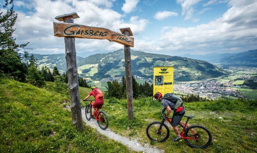 Zwei Mountainbiker am hölzernen Eingangstor des Gaisberg Trails mit Blick auf ein Tal.