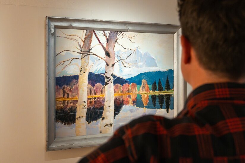 Ein Mann betrachtet ein gerahmtes Gemälde von Alfons Walde mit Birken, einem See und Bergen im Hintergrund.