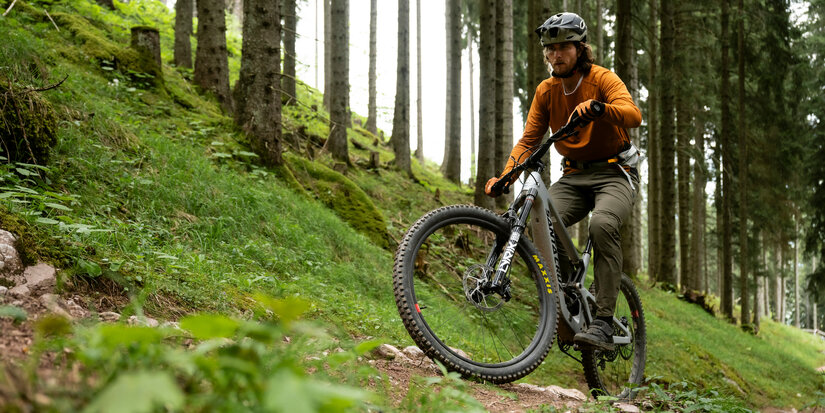 In Kitzbühel, a cyclist wearing an orange jersey rides confidently along a rocky path through a dense forest.