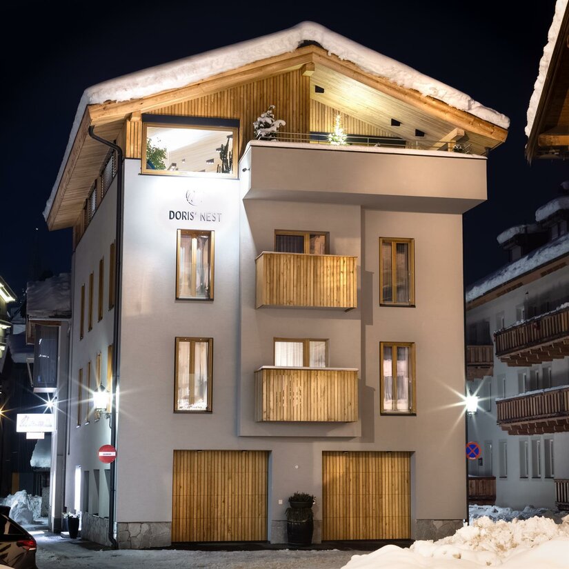 Unterkunft Doris' Nest Beleuchtetes, mehrstöckiges Gebäude mit Holzfassadenelementen und schneebedecktem Dach bei Nacht.