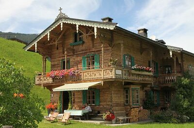 Landhaus Kainzern im Sommer Zweistöckiges Holzhaus im alpinen Stil mit Blumenbalkonen und grünen Fensterläden vor einer Wiese.