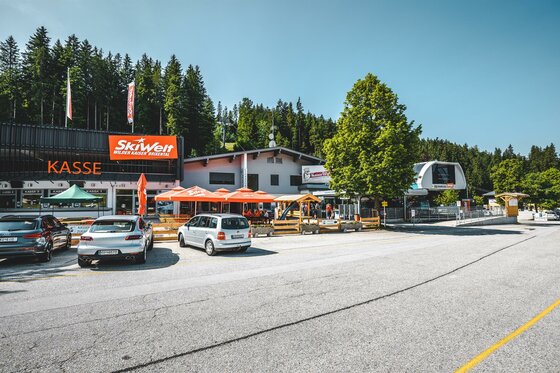 Eine belebte Bergstation mit einem Kassenbereich und zahlreichen Fahrzeugen. Die Umgebung ist grün und von Bäumen umgeben, ideal für Outdoor-Aktivitäten.