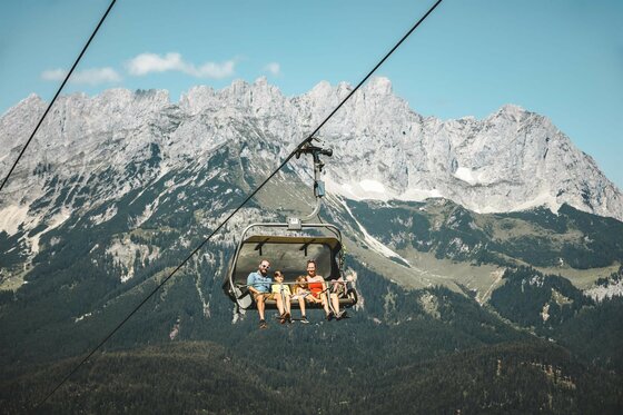 Going_PonyAlm_Wilder Kaiser (2) Eine Seilbahn mit vier Personen fährt über eine wunderschöne Berglandschaft. Im Hintergrund sind majestätische Berge und ein blauer Himmel zu sehen.