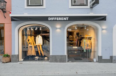 Außenansicht eines Modegeschäfts mit zwei bogenförmigen Schaufenstern und schwarzer Markise.