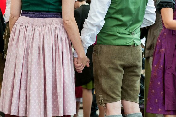 Ein Paar hält Händchen. Die Person links trägt einen Rock, während die Person rechts eine traditionelle Tracht mit kurzen Hosen trägt.