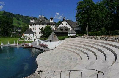 Ein malerisches Schloss mit einem grünen Umfeld und blauen Himmel. Im Vordergrund gibt es eine elegante Treppe, die zu einem Teich führt.