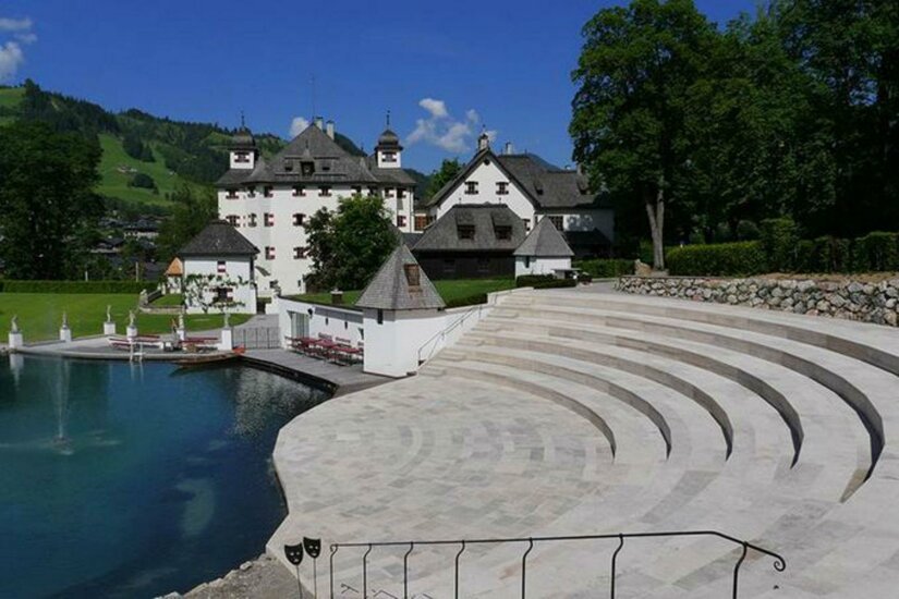 Kaps Ein malerisches Schloss mit einem grünen Umfeld und blauen Himmel. Im Vordergrund gibt es eine elegante Treppe, die zu einem Teich führt.