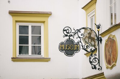 Museum Ein elegantes Museums-Schild hängt an einer weißen Wand. Daneben befindet sich ein Fenster mit gelben Fensterrahmen.