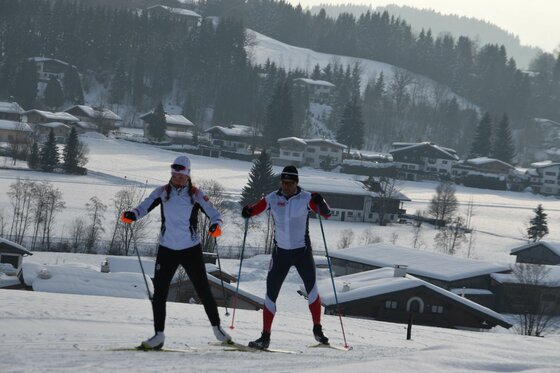 Zwei Personen beim Skilanglauf auf einer verschneiten Loipe vor einer dörflichen Kulisse im Winter.