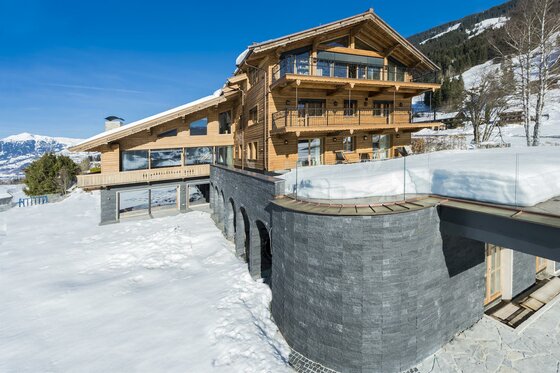 Mehrstöckiges Holzchalet mit Steinfassade in einer verschneiten Berglandschaft unter blauem Himmel.