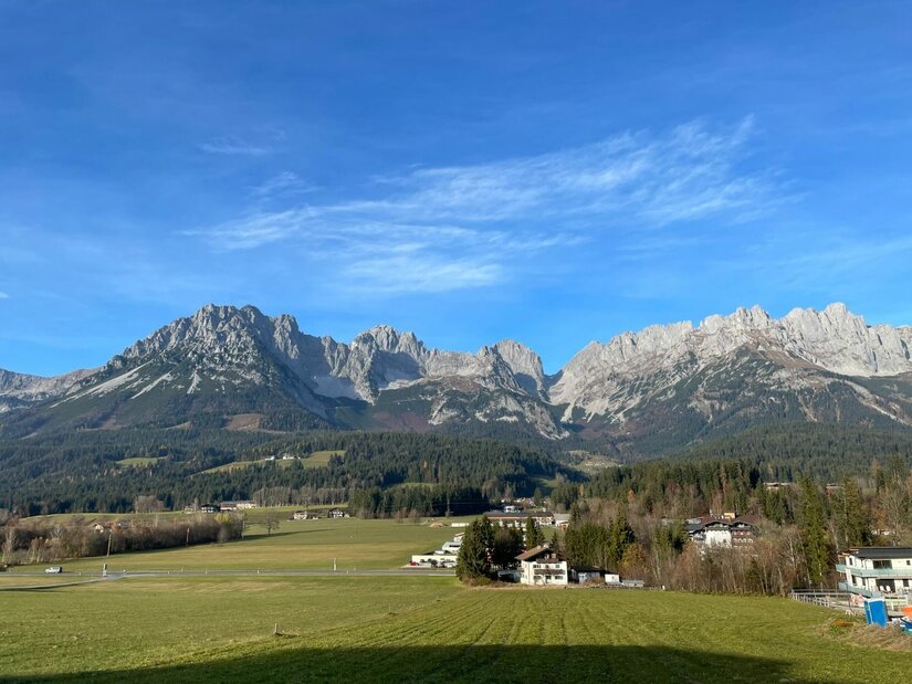 Panorama Wilder Kaiser Blick auf das Gebirgsmassiv des Wilden Kaisers über grüne Wiesen und vereinzelte Häuser unter blauem Himmel.