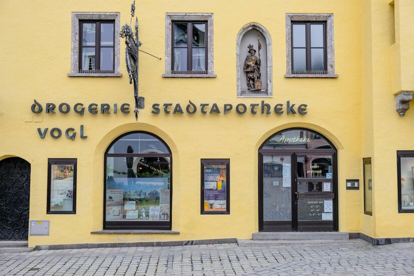 Stadtapotheke Vogl Gelbe Fassade der Stadtapotheke Vogl mit Rundbogenfenstern, einer Heiligenfigur und historischer Beschriftung.