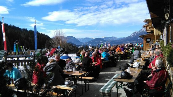 Going_Hollenauer Kreuz_Terasse_Sommer_2022_3 Eine belebte Terrasse mit vielen Gästen, die die Sonne genießen. Im Hintergrund sind Berge und ein klarer Himmel zu sehen.