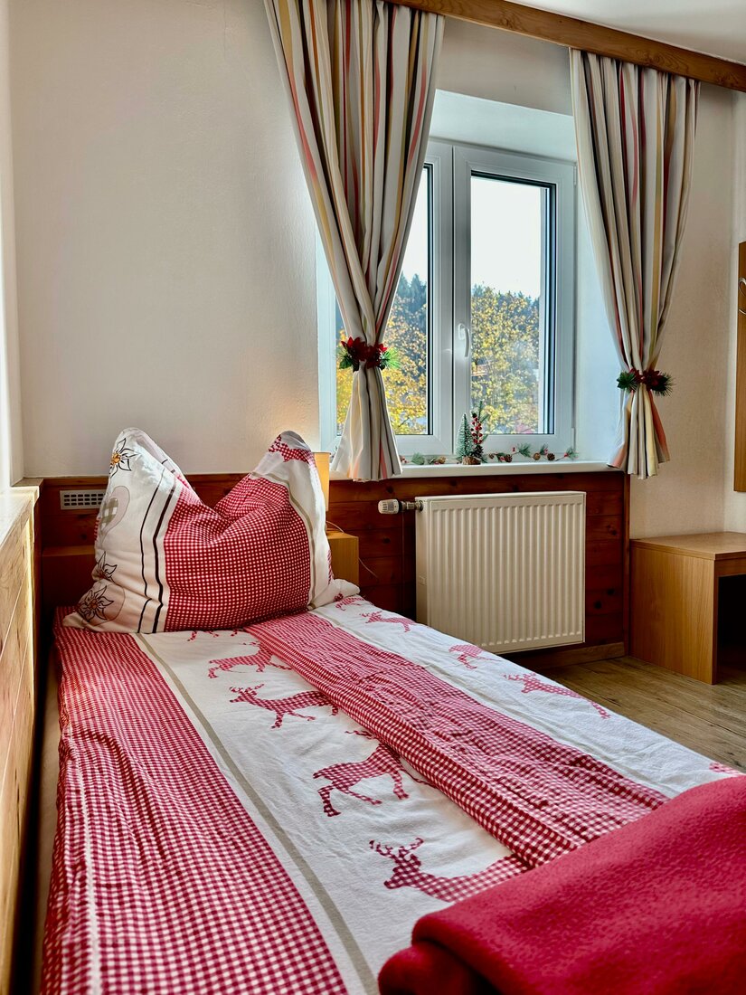 Einzelbett mit rot-weißer Bettwäsche vor einem Fenster mit Vorhängen.