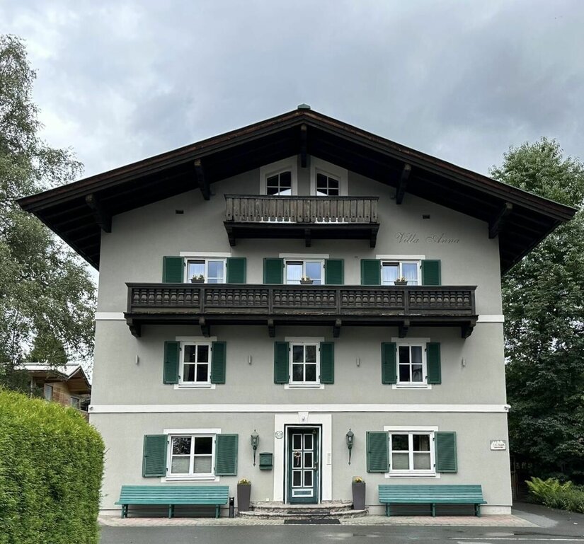 Vorderansicht der Villa Anna mit grauer Fassade, grünen Fensterläden und Holzbalkonen unter bewölktem Himmel.