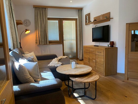 Ein gemütliches Wohnzimmer mit einem grauen Sofa, einem runden Tisch und einem Fernseher an der Wand. Das Licht strömt durch die Fenster und sorgt für eine warme Atmosphäre.