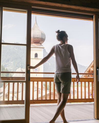 Eine Person steht auf einem Balkon und blickt auf eine malerische Berglandschaft mit einer Kirchturmspitze im Hintergrund.