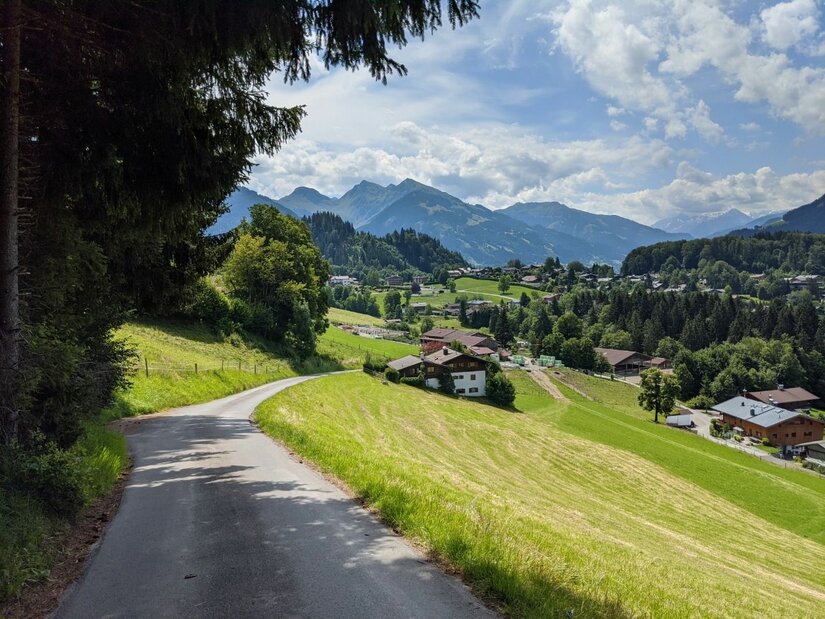 Bichlalmlift-Hornbahn Wanderweg Asphaltierter Weg führt an grünen Wiesen und Häusern vorbei auf ein Bergpanorama zu.