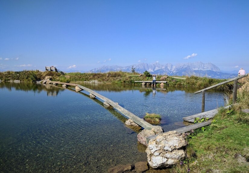 Holzsteg am Bergsee Holzsteg führt über einen klaren Bergsee vor einem Gebirgspanorama unter blauem Himmel.