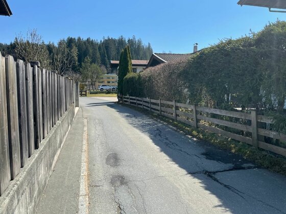 Wegverlauf in Rettenberg Asphaltierter Weg zwischen Holzzäunen und Hecken in einer Siedlung vor bewaldetem Hintergrund.
