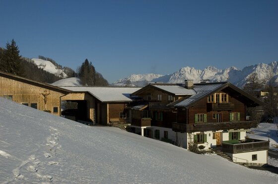 Ferienhof Oberstaudach im Winter Alpiner Ferienhof aus Holz und Stein in einer verschneiten Berglandschaft unter blauem Himmel.