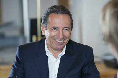 Christian Krassnigg Immobilien Ein lächelnder Mann trägt einen dunkelblauen Anzug. Er sitzt an einem Tisch und scheint in einem Gespräch zu sein.