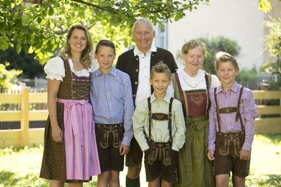 Familie Schmidinger in Tracht Sechs Personen in traditioneller Tracht stehen lächelnd in einem Garten vor einem Holzzaun.