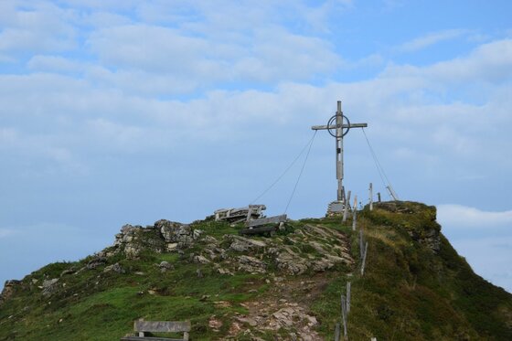 Gipfelkreuz am Kitzbüheler Horn Ein hölzernes Gipfelkreuz auf einem felsigen, grasbewachsenen Berggipfel unter leicht bewölktem Himmel.
