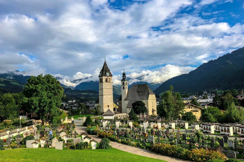 Kirche mit Friedhofsanlage Blick auf eine Kirche mit zwei Türmen und einen angrenzenden Friedhof vor einer Bergkulisse.