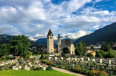 Blick auf eine Kirche mit zwei Türmen und einen angrenzenden Friedhof vor einer Bergkulisse.