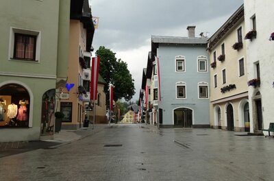 Fußgängerzone in Kitzbühel Gepflasterte Fußgängerzone in Kitzbühel mit bunten Häuserfassaden und rot-weißen Fahnen unter bewölktem Himmel.