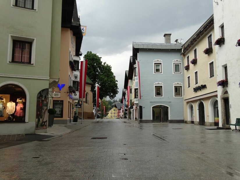 Fußgängerzone in Kitzbühel Gepflasterte Fußgängerzone in Kitzbühel mit bunten Häuserfassaden und rot-weißen Fahnen unter bewölktem Himmel.
