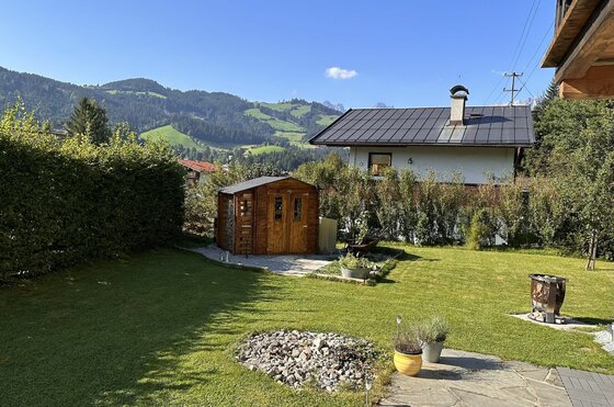 Garten mit Rasenfläche, Holzschuppen und Wohnhaus vor bewaldeten Bergen.